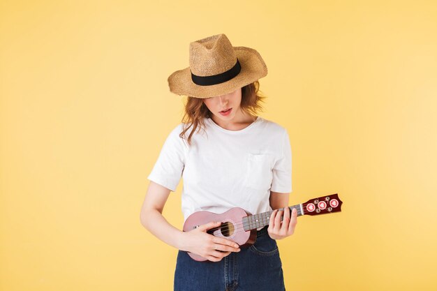 小さなギターと立ってピンクの背景の上でそれで遊んで麦わら帽子のきれいな女性の肖像画