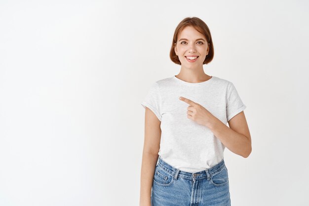 Портрет красивой дамы в футболке, указывающей влево и улыбающейся, рекомендующей продукт, рекламная компания, стоящая у белой стены