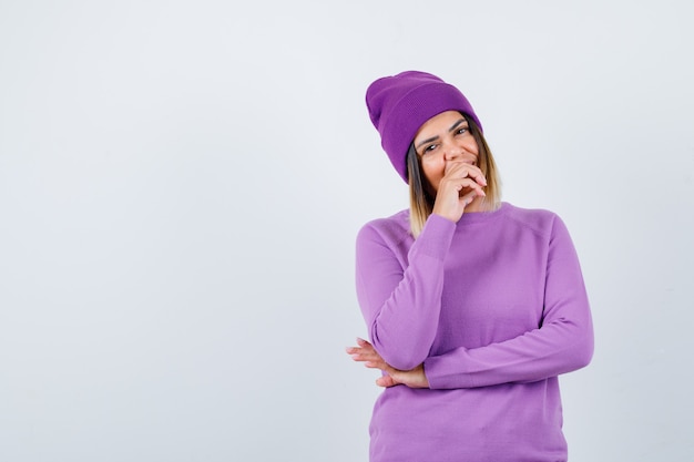 Portrait of pretty lady with hand on mouth in sweater, beanie and looking cheerful front view