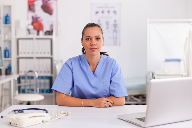 Portrait of pretty medical nurse smiling at camera in hospital office wearing blue uniform. Healthcare practitioner sitting at desk using computer in modern clinic looking at monitor, medicine.