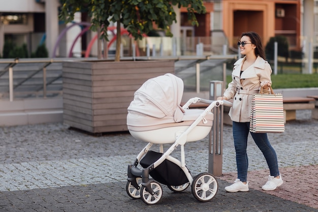 Free photo portrait of pretty mother with shopping package walking with baby carriage incity center