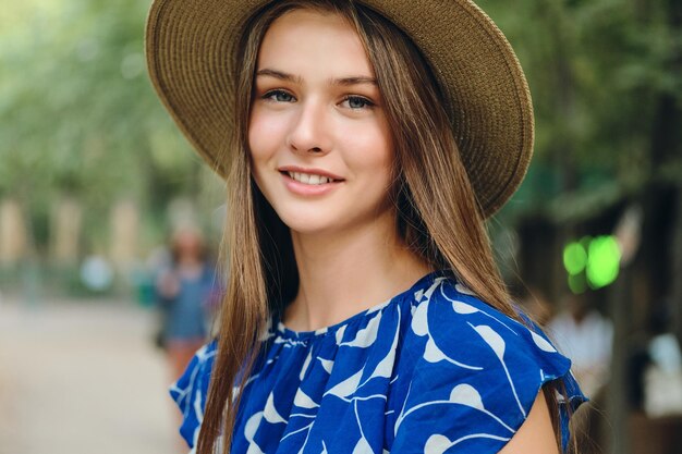 Portrait of pretty smiling girl in blue dress and hat dreamily looking in camera in city park