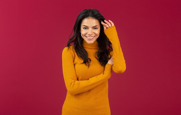 Портрет довольно улыбающейся стильной брюнетки латиноамериканского происхождения в желтом осеннем зимнем модном платье-свитере, позирующей изолированно на красной стене