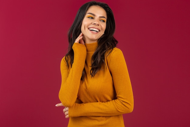 Portrait of pretty smiling stylish brunette hispanic woman in yellow autumn winter fashion dress sweater posing isolated on red wall 