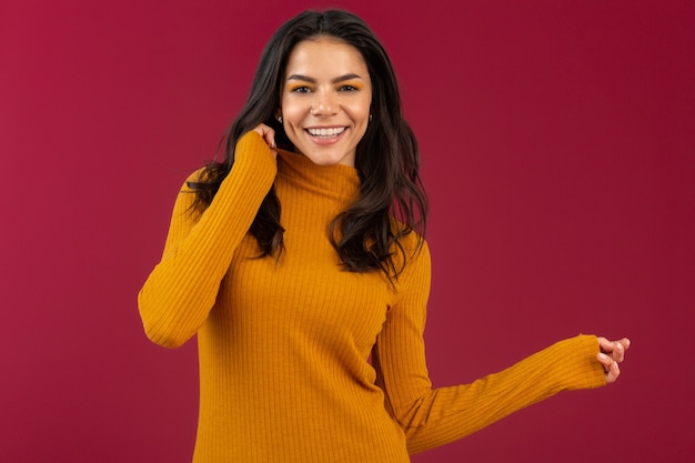 Foto gratuita ritratto della donna ispanica castana alla moda abbastanza sorridente in maglione giallo del vestito da modo di inverno di autunno che posa isolata sulla parete rossa