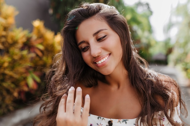 Portrait of pretty tanned woman with long curly hair smiling against wall of trees