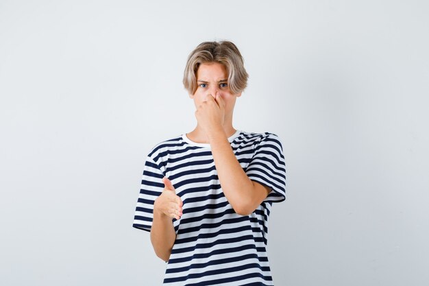 Portrait of pretty teen boy smelling something awful, pinching nose, showing stop sign in striped t-shirt and looking disgusted front view