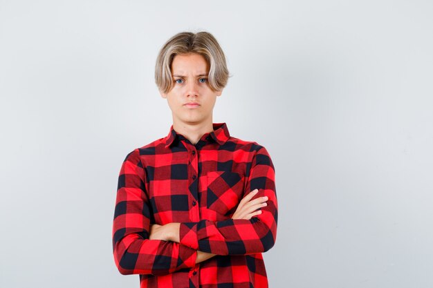 Портрет симпатичного мальчика-подростка, стоящего со скрещенными руками в клетчатой рубашке и недовольного вида спереди