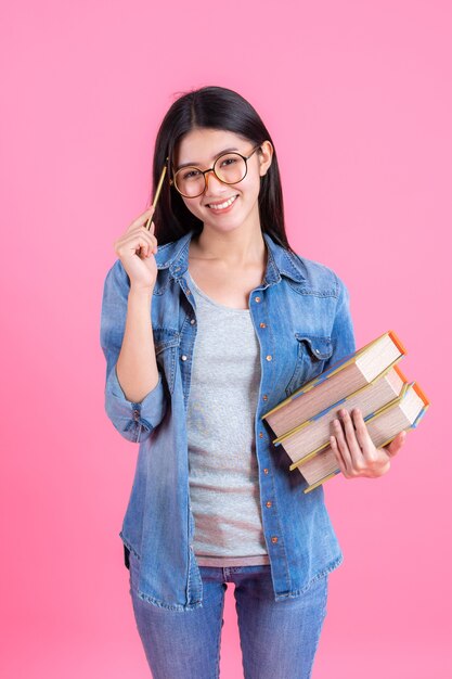 Portrait pretty teenage female holding books in her arm and using pencil on pink, education concept