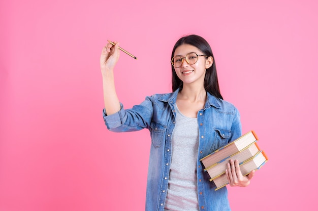 Portrait pretty teenage female holding books in her arm and using pencil on pink, education concept