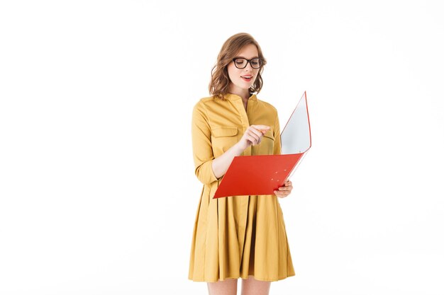 Портрет довольно задумчивой дамы в очках и желтом платье, стоящей с красной папкой в руке на белом фоне изолированы