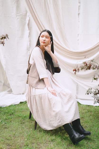 Portrait of pretty woman at garden, sitting on a chair and looking in cream color dress during daytime.