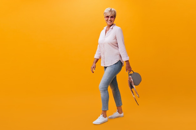 Портрет красивой женщины в джинсах и рубашке, позирующей с сумкой на оранжевом фоне