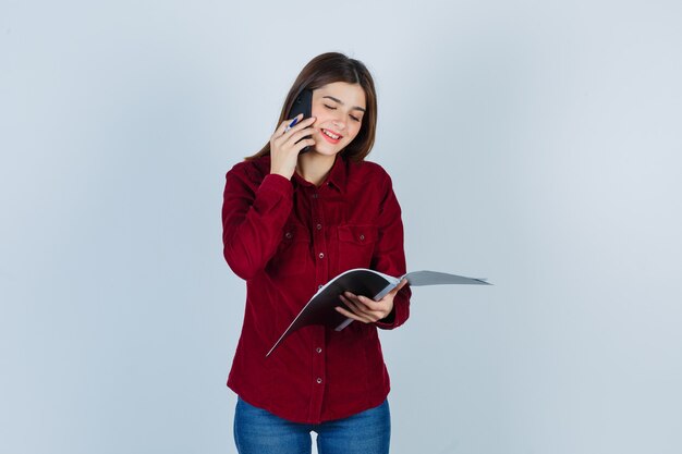 Portrait of pretty woman keeping pen, folder, talking on smartphone while smiling in burgundy blouse and looking cheerful