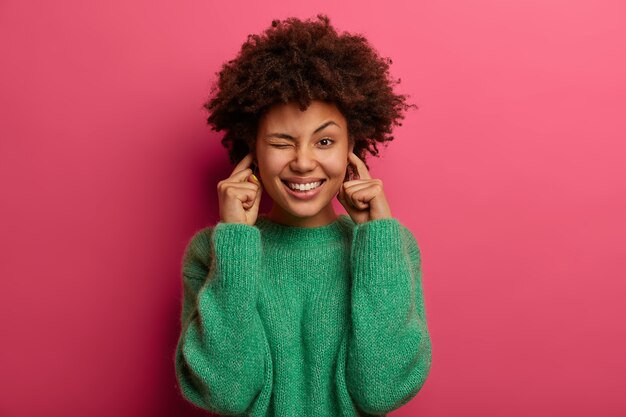 Portrait of pretty woman plugs ears and winks eyes, smiles broadly, ignores unpleasant noisy sound, wears green sweater, poses against pink wall, has happy expression. Turn off volume.