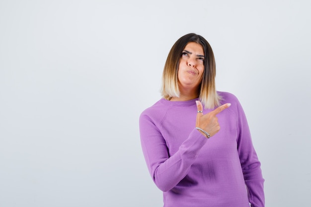 Portrait of pretty woman pointing at upper right corner, looking up in purple sweater and looking pensive front view