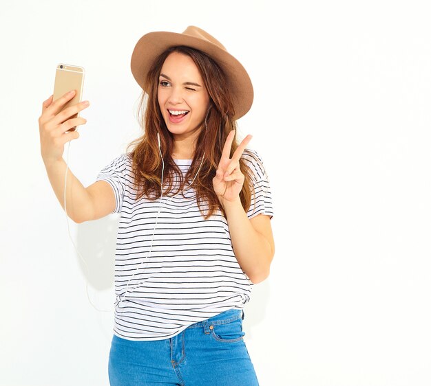 Portrait of a pretty woman in summer hipster clothes taking a selfie isolated on white wall. Winking and showing peace sign