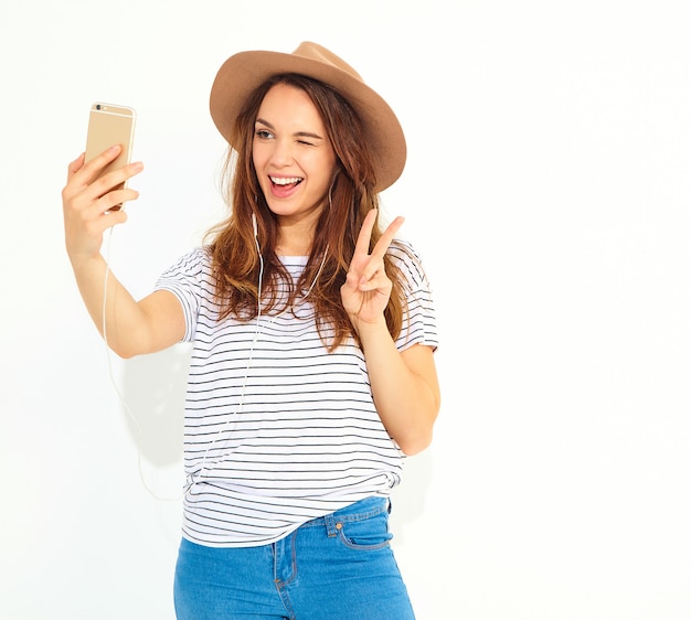 Portrait of a pretty woman in summer hipster clothes taking a selfie isolated on white wall. Winking and showing peace sign