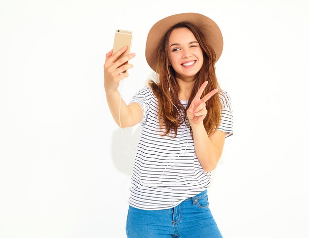 Portrait of a pretty woman in summer hipster clothes taking a selfie isolated on white wall. Winking and showing peace sign