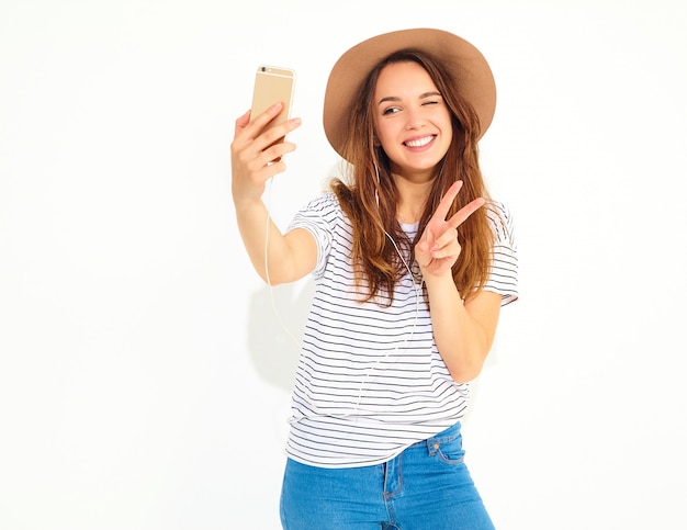 Portrait of a pretty woman in summer hipster clothes taking a selfie isolated on white wall. Winking and showing peace sign