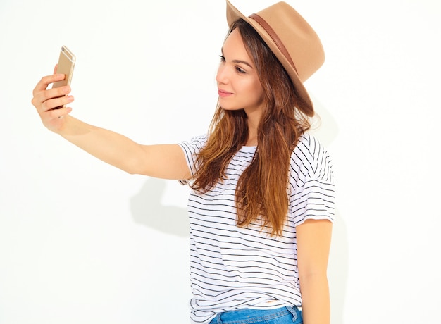 Portrait of a pretty woman in summer hipster clothes taking a selfie isolated on white wall