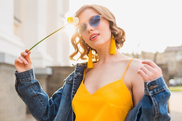 Portrait of pretty woman wearing heart sunglasses holding flower against sun, sunny summer day, stylish apparel, fashion trend, blue jeans jacket, yellow dress, elegant, hister, earrings