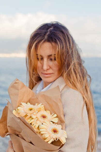 Портрет симпатичной женщины с цветами, стоящими и выглядящими счастливыми на побережье в течение дня.