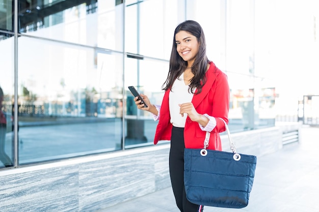 Portrait of pretty young businesswoman holding smartphone and purse while walking in city