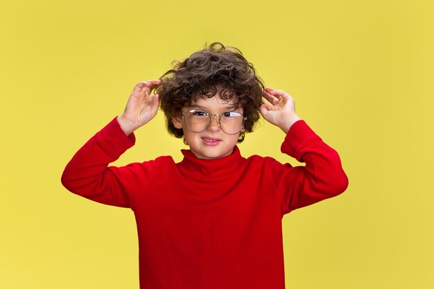 Portrait of pretty young curly boy in red wear on yellow studio background. Childhood, expression, education, fun concept.