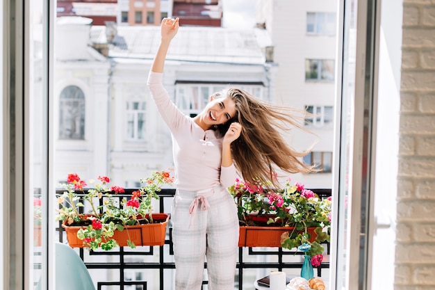 Portrait pretty young girl in pajamas having fun on balcony in city. She moves, raises hands. Her long hair flying and she smiling .