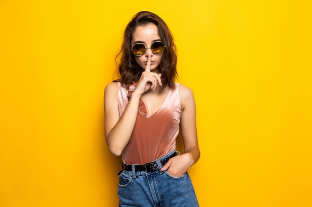 Portrait of a pretty young girl showing silence gesture and winking isolated over yellow 