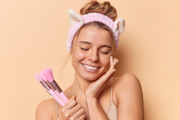 Portrait of pretty young woman touches face gently keeps eyes closed holds cosmetic brushes keeps eyes closed wears headband smiles toothily isolated over beige background. Beauty time concept