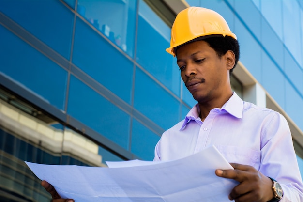 Portrait of professional architect in helmet looking at blue prints outside modern building. Engineer and architect concept.