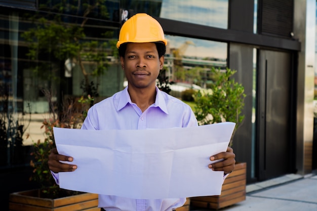 Portrait of professional architect in helmet looking at blue prints outside modern building. Engineer and architect concept.