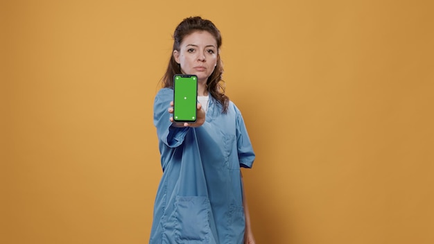 Portrait of professional medic holding smartphone and texting and showing green screen mockup design at camera in studio. Doctor in hospital uniform using touchscreen device presenting copy space.