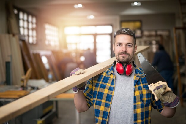 Portrait of professional middle aged carpenter with wood plank and tools standing in his woodworking workshop