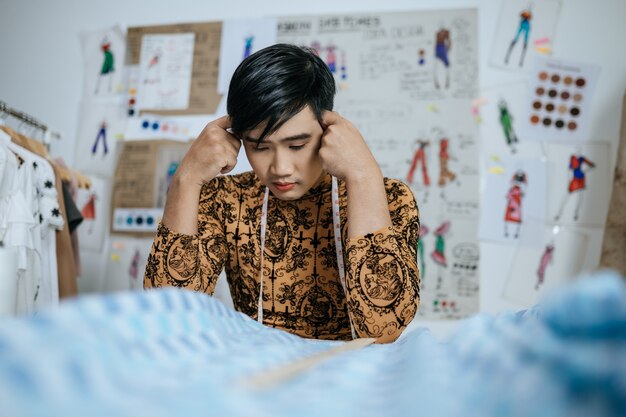 Portrait of Professional stressed asian young male tailor with measuring tape on neck touching head with hands in studio.