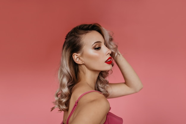 Portrait in profile in studio of beautiful young blonde with curly hair and brightly painted pink lips, posing for camera showing her tender feminine shoulders