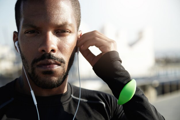 Portrait of purposive fitness trainer preparing himself for tough workout. With serious face, tracker, headphones in ears African American athlete is determined to challenge himself in sport.