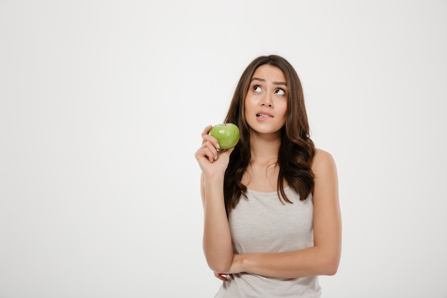 Портрет озадаченная женщина, глядя вверх, держа зеленое свежее яблоко, думая о здоровой пищи, изолированных на белый