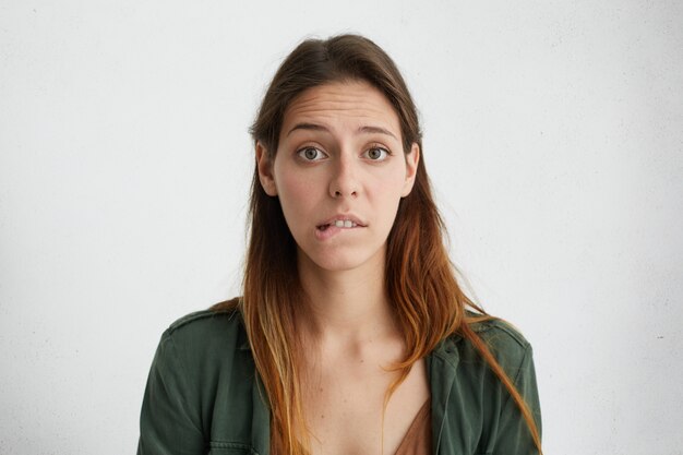 portrait of puzzled woman with long face and straight dyed hair wearing green jacket looking with big opened eyes biting her lower lip having some doubts and uncertainty