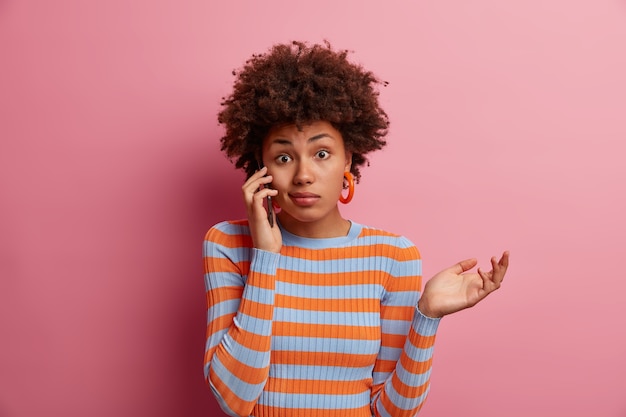 Portrait of questioned Afro American woman has telephone conversation and hesitates with answer, makes weighing gesture and raises palm doubtfully, being unaware and has no idea, not sure about