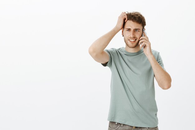 Portrait of questioned cute fair-haired man with bristle, holding smartphone near ear and scratching head while trying answer question, being unsure and doubtfu