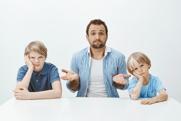 退屈で動揺している息子と一緒にテーブルに座って、上げられた手のひらで肩をすくめて、一人で男の子を育てる方法が無知である疑わしい見栄えの良いヨーロッパの父の肖像