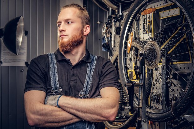 自転車の部品とホイールを背景にしたワークショップでの赤い頭のひげを生やした自転車整備士の肖像画。