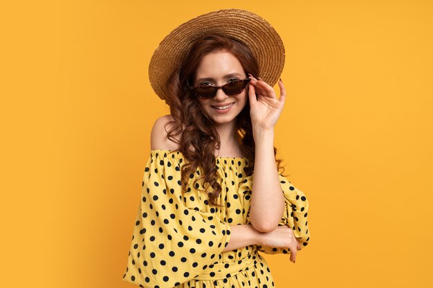 портрет рыжей женщины в соломенной шляпе и стильных солнцезащитных очках, позирующих на желтом в летнем платье.
