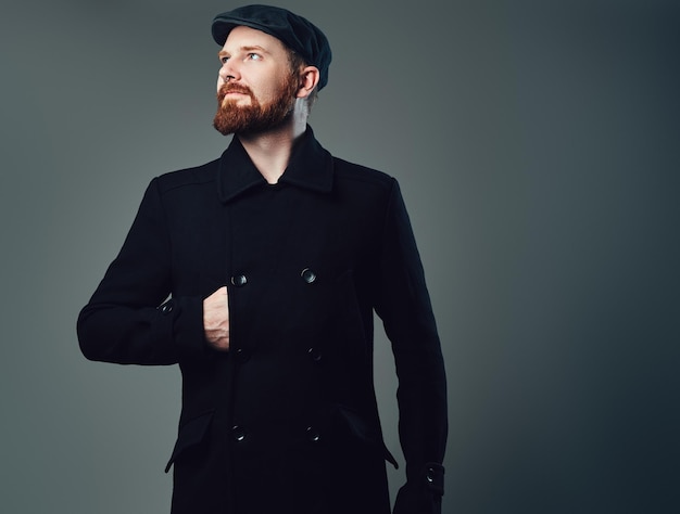 Portrait of Redbeard elegant male dressed in a tweed flat cap and a black jacket over grey background.