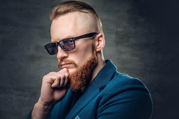 Portrait of redhead bearded hipster male dressed in a blue jacket and sunglasses over grey background.