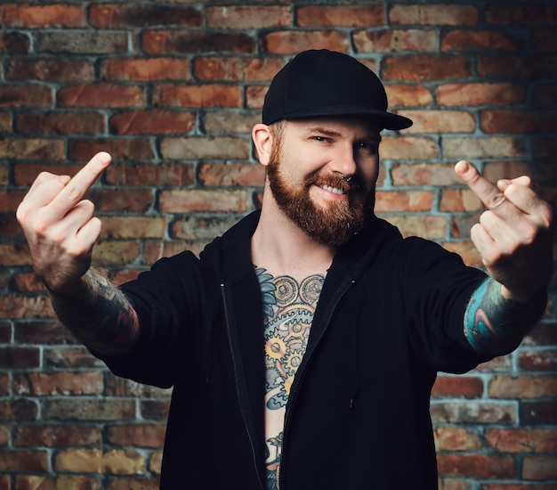 Portrait of redhead bearded male with tattoos on arms and chest, dressed in ablack sports jacket over the wall of a brick.