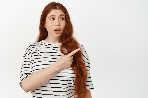 Portrait of redhead curvy girl with surprised face, showing something strange, pointing finger right and looking at promo sale with clueless face, asking question, white background.